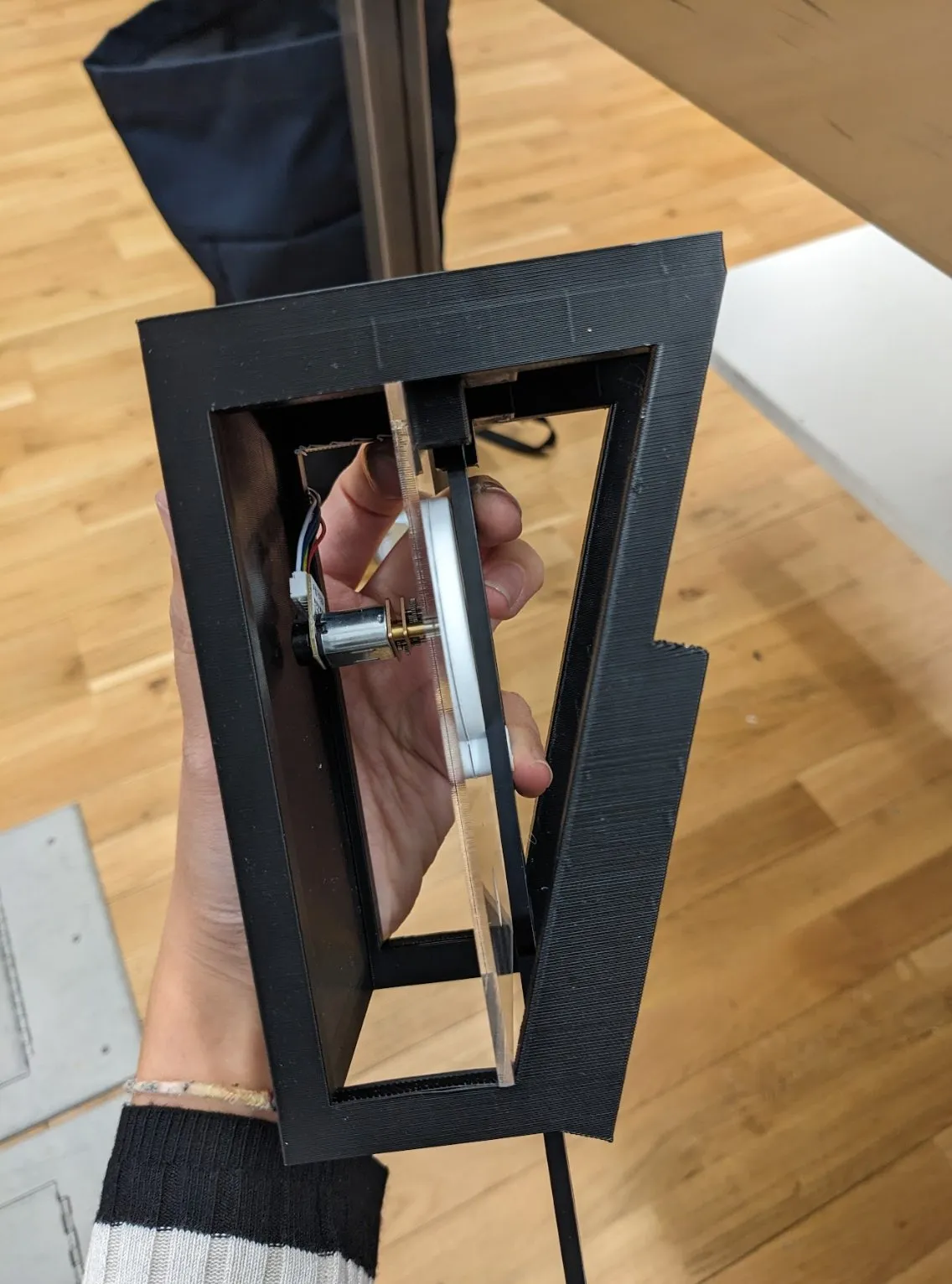 The black frame held over a wooden floor with the pendulum arm and lipstick mounted inside it.