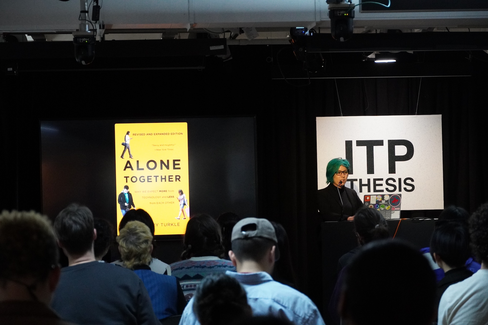 "A photograph of a woman with green hair presenting a slide with a picture of a book with the title "Alone Together"