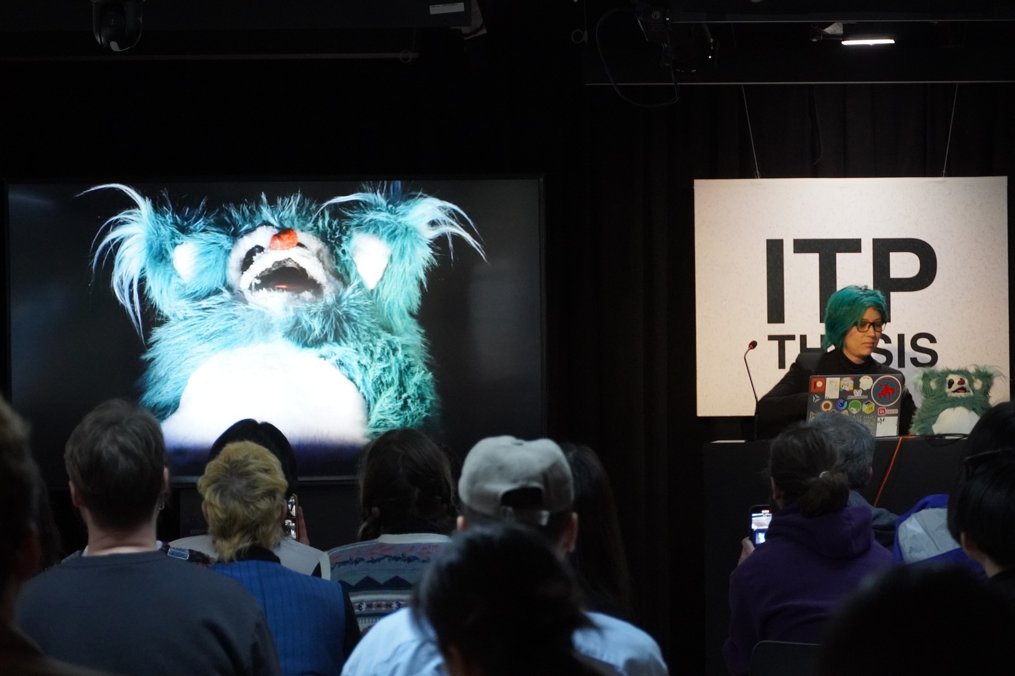 "A photograph of a woman with green hair presenting a slide with a giant photograph of a fluffy teal stuffed animal with its mouth open with ominous lighting.