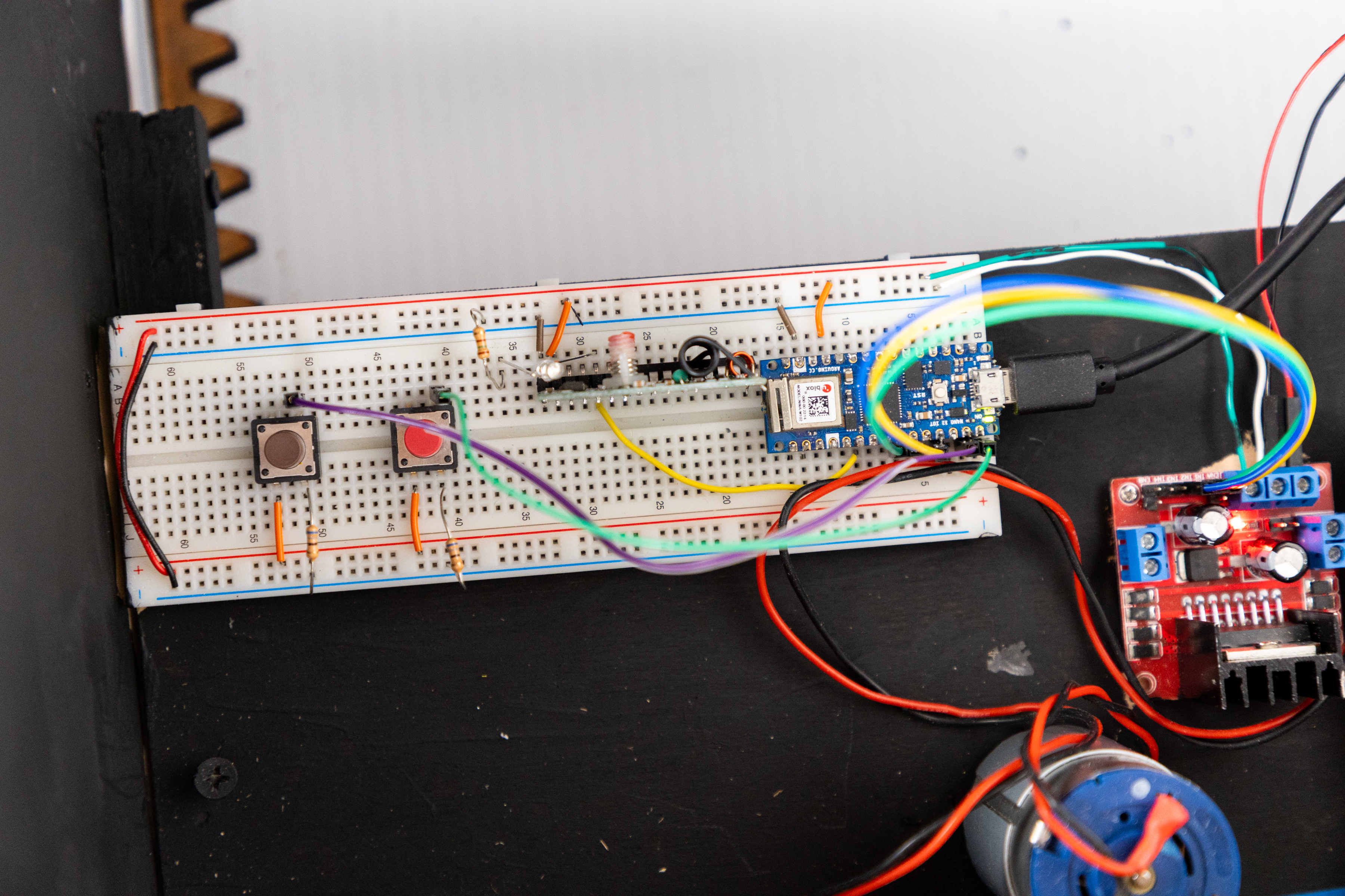 The image shows a close-up of a breadboard setup for an electronic project. It features two push buttons on the left side, connected with various colored wires. A small microcontroller board is positioned in the center, with multiple wires extending from it. To the right, there is a red circuit board with two adjustable knobs and an LED indicator. Below the breadboard, a small motor is visible, connected by wires. The background is a dark surface, emphasizing the components of the circuit.