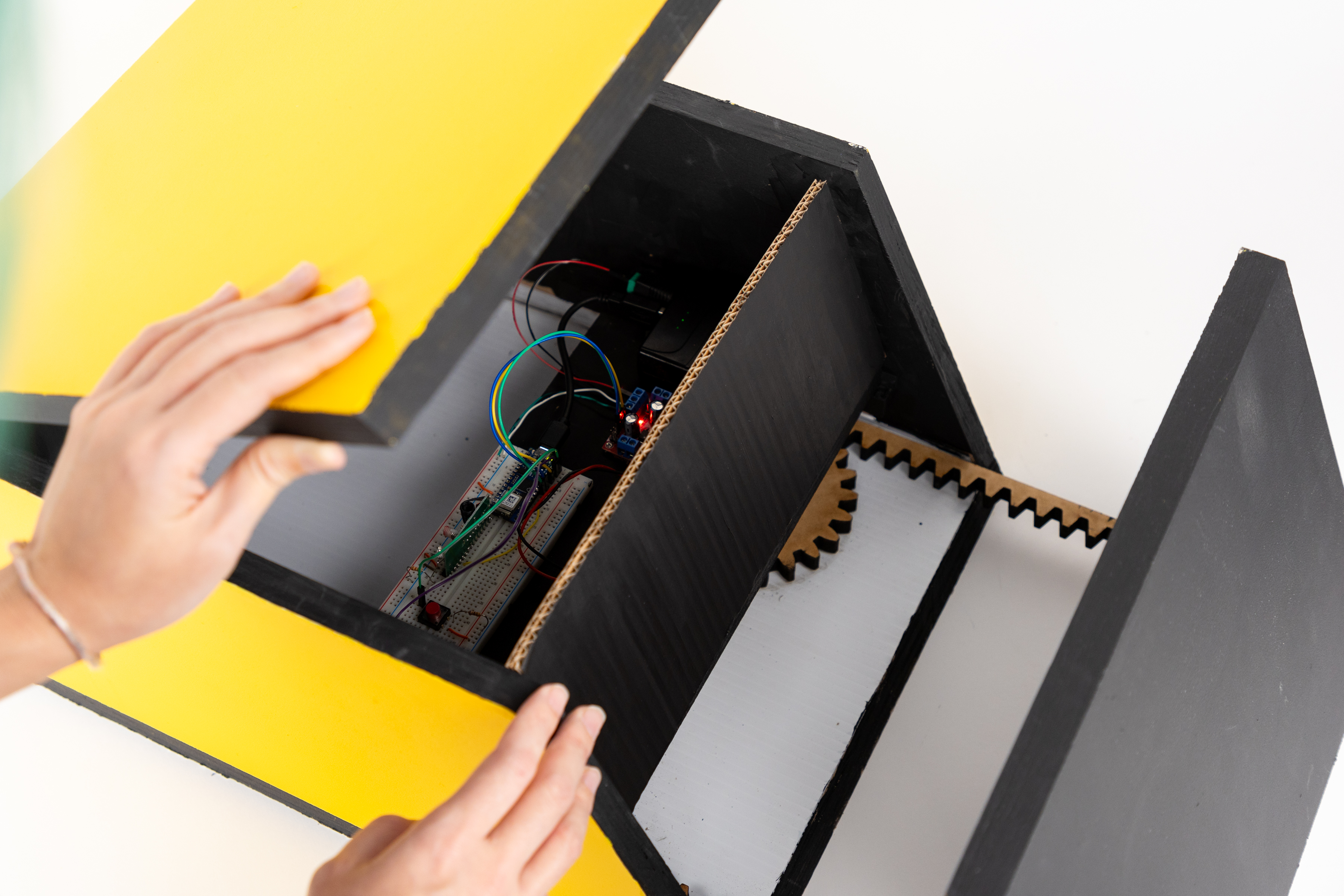 The image shows a person’s hands opening a box with a yellow lid and a black body. Inside the box, there are various electronic components, including a circuit board with wires connected to it. A red LED light is visible, indicating power. The interior also features a gear mechanism, with a large wooden gear positioned on the bottom. The overall setup suggests a project involving electronics and mechanical parts, highlighting the functional design of the box.