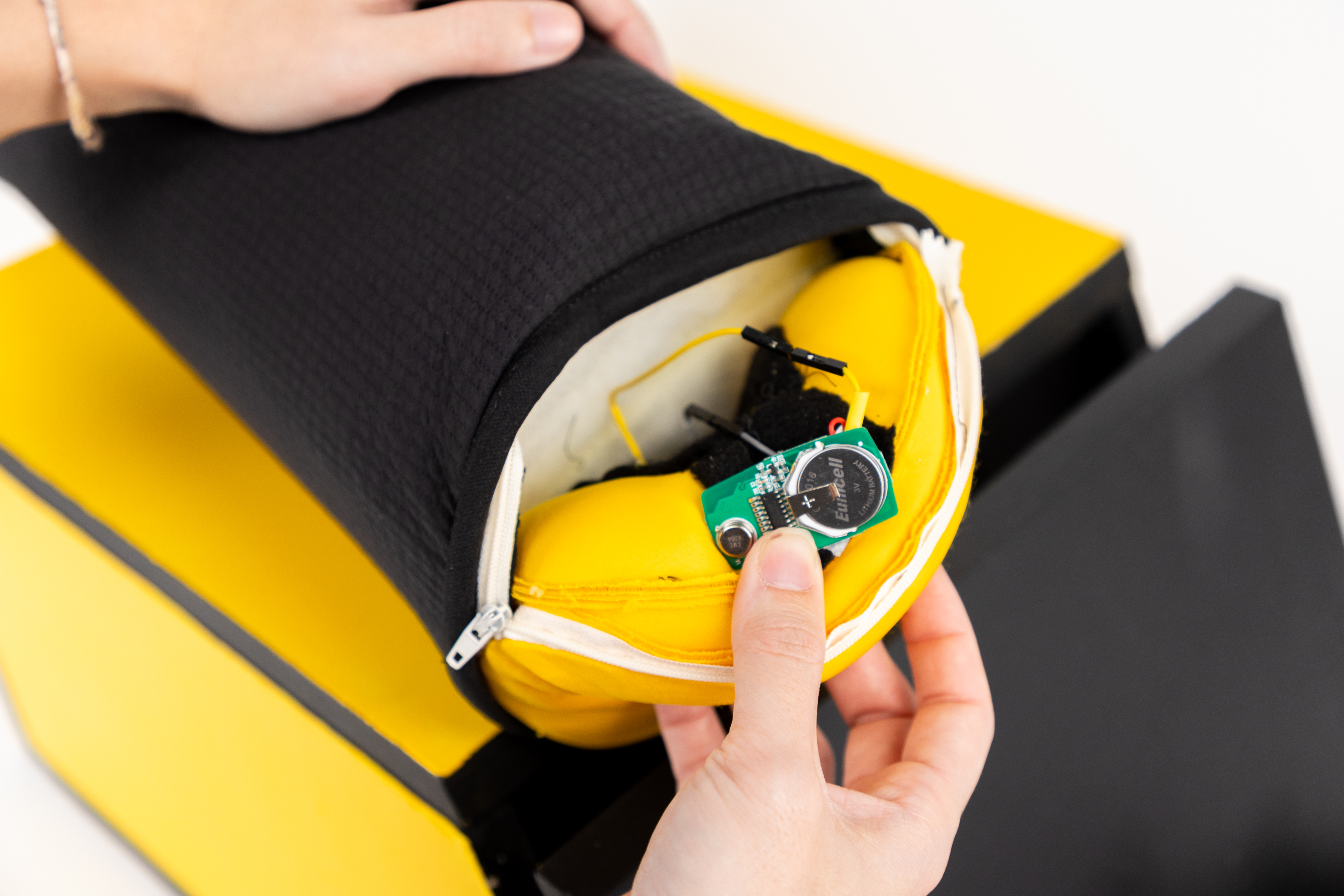 The image shows a close-up of a hand holding a cylindrical object with a black exterior and a yellow interior. The zipper is partially open, revealing electronic components inside. A small green circuit board with a battery is visible, along with wires connected to it. The background features a yellow and black structure, emphasizing the design of the object. The focus is on the hand interacting with the zipper and the internal components, highlighting the functional aspect of the item.