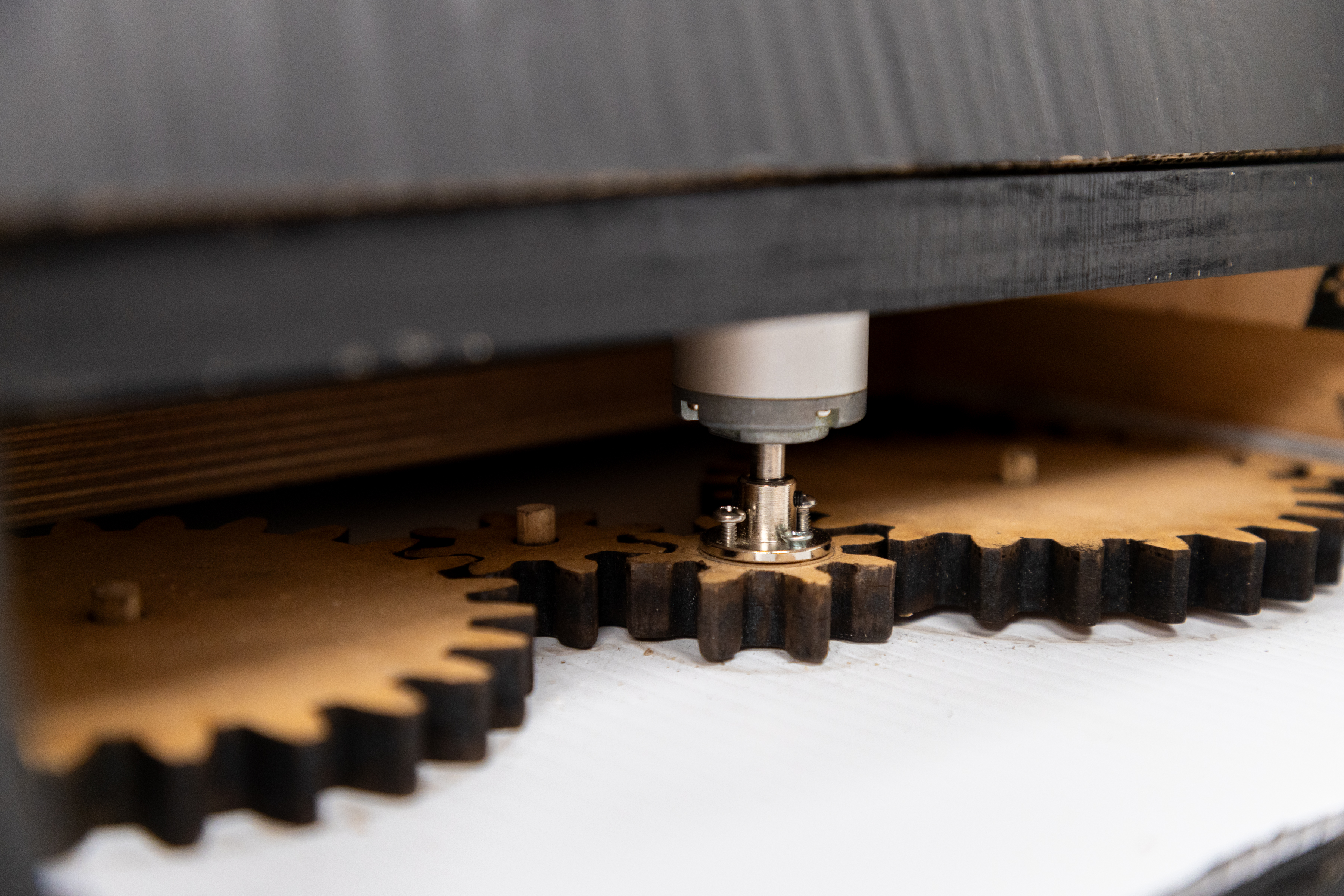 The image shows a close-up view of a mechanical setup involving gears. A motor is positioned above two large, wooden gears with distinct teeth. The motor has a metallic shaft that is aligned with the center of one of the gears. The gears are placed on a flat surface, and the background is slightly blurred, focusing attention on the mechanical components. The overall scene suggests a working environment, possibly related to a project involving gear mechanisms or machinery.