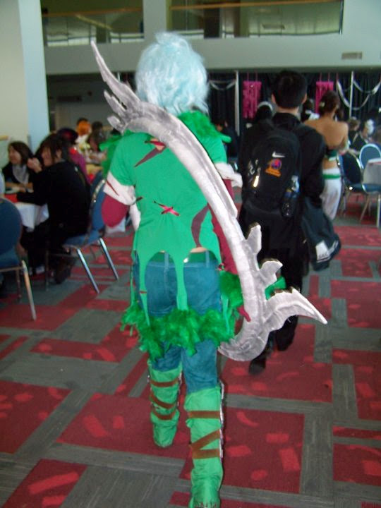 Photograph of a girl faced the other way in a green feather outfit and blue hair resting a giant ring with blades on her shoulder